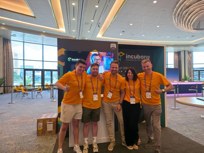 Five members of the Incubane team in orange Incubane shirts, arms around each other in front of their branded stand at Workday DevCon.